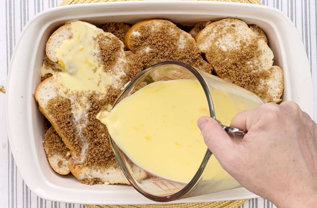 Pouring an egg mixture over a French toast casserole in a dish.