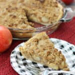 A slice of apple crumb pie in front of the whole pie with some apples