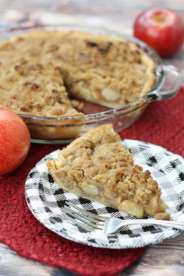 A slice of apple crumb pie in front of the whole pie with some apples