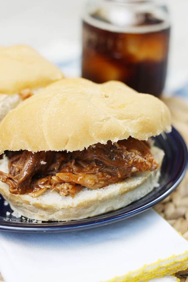 A close up pic of a root beer pulled pork sandwich on a blue plate in front of a glass of root beer
