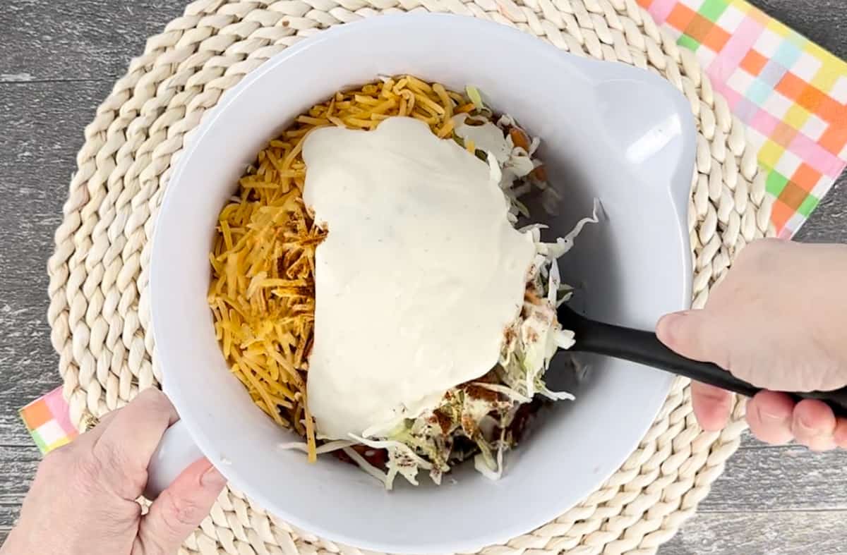 Adding ranch dressing to ingredients in a white bowl with a spatula.