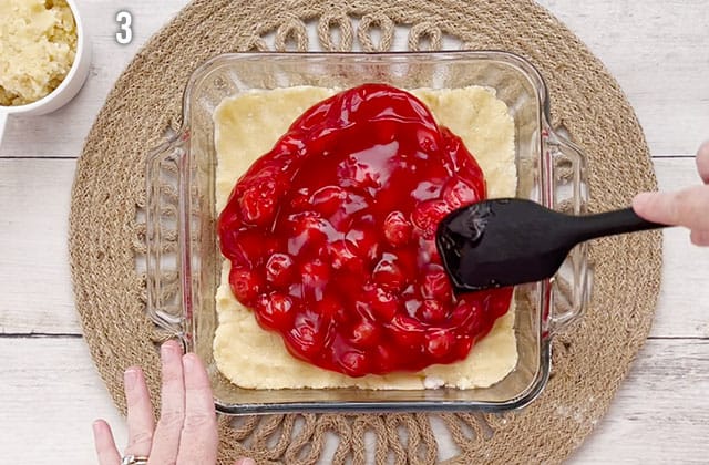 Spreading cherry pie filling over the sugar cookie dough crust in a baking dish