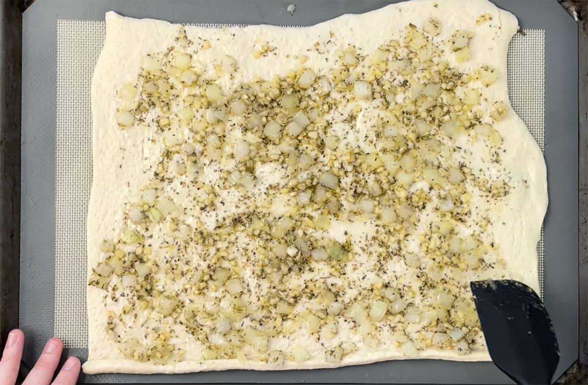 Spreading an herb mixture over pizza dough on a cookie sheet.
