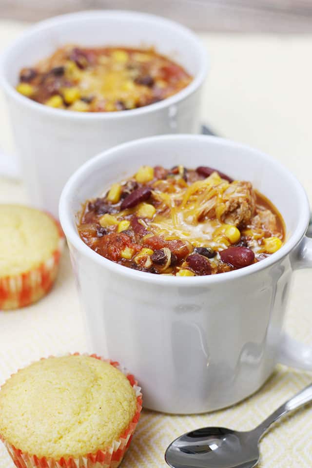 Two white soup mugs filled with crockpot chicken chili with cornbread muffins next to them