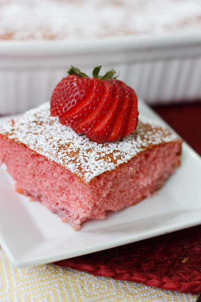 Strawberry jello cake on a white square plate topped with powdered sugar and sliced strawberry