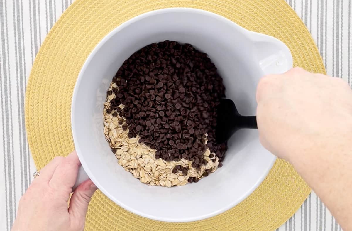 Stirring chocolate chips and old fashioned oats in a white mixing bowl.