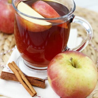 A glass mug of apple tea next to cinnamon sticks.