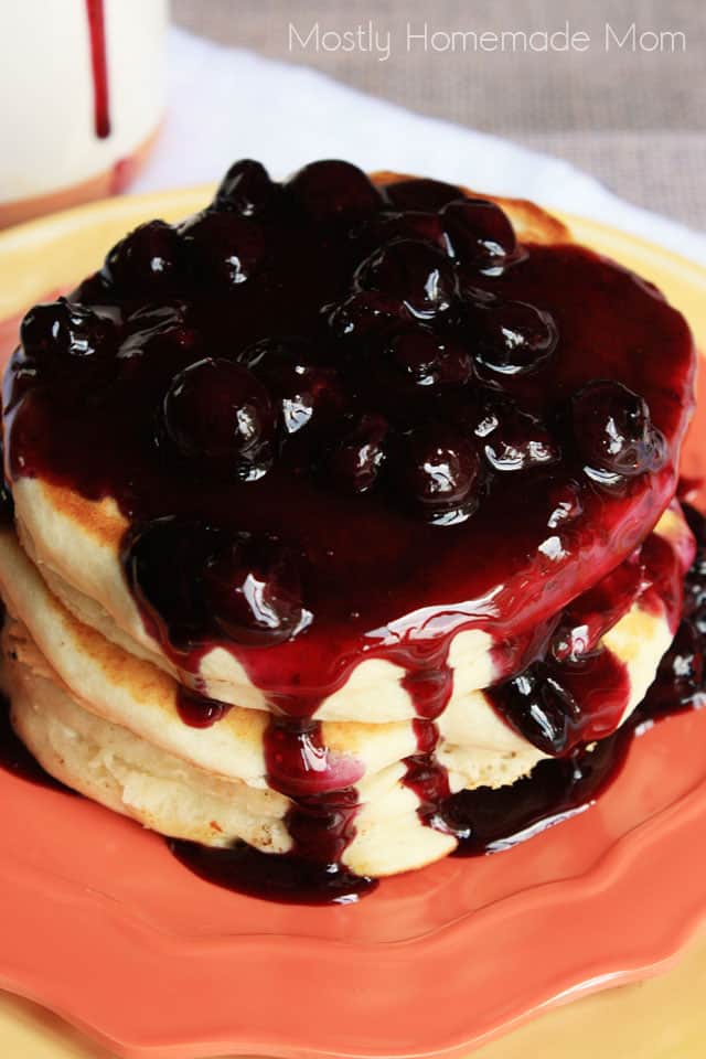 A stack of three pancakes on an orange plate topped with homemade blueberry syrup