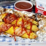 Corn on the cob wrapped in bacon and drizzled with bbq sauce on foil in a tray
