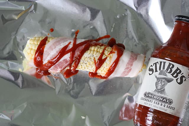 Ear of corn wrapped in bacon and drizzled with bbq sauce on foil