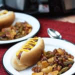 Crockpot baked beans on white plates next to hot dogs