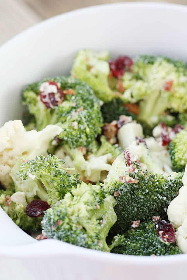 A close up picture of broccoli cauliflower salad in a white mixing bowl being served