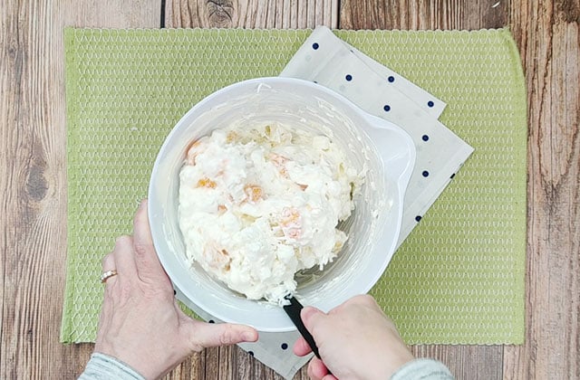 Mixing ambrosia salad ingredients in a white bowl with a black spatula